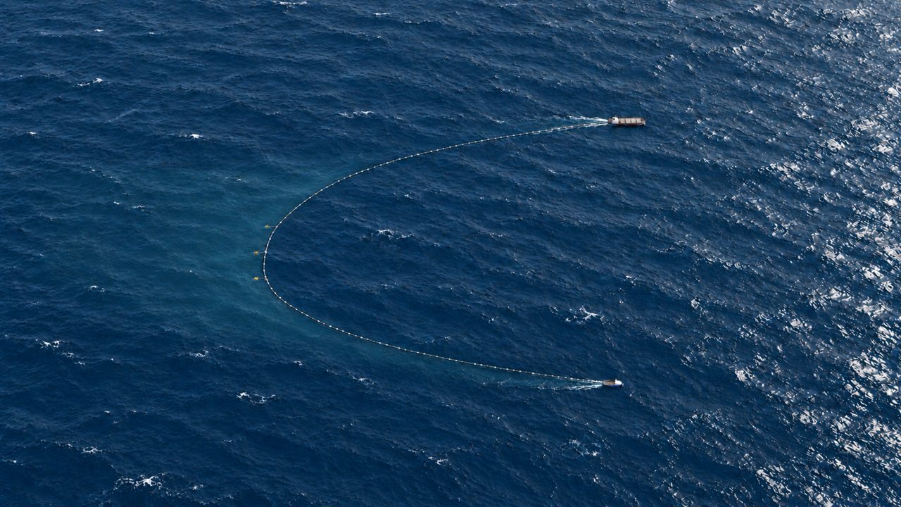 Aerial concept view of the vessels advancing to the right, with the floating hose and a faint, broad treated-water area extending behind them to the left.